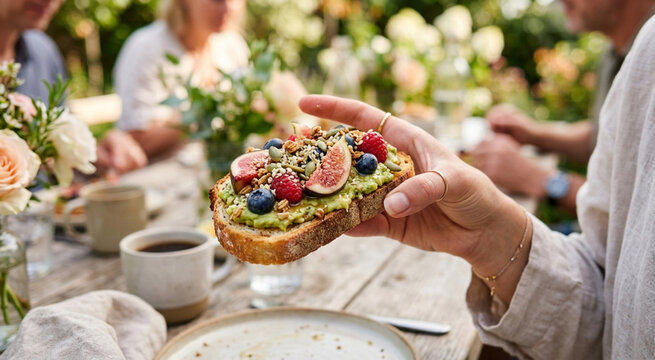 Close-up of hand holding healthy avocado toast topped with fresh figs, berries, and seeds at an outdoor garden brunch.