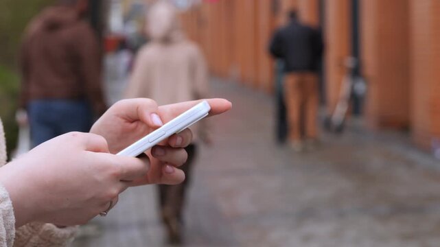 Close-up of hands holding smartphone while walking outdoors. Fingers scroll screen. Background shows blurred pedestrians on sidewalk. Person appears focused on device. Ideal for tech, urban life