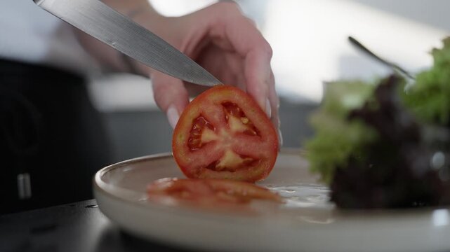 A closeup shot of hands using a sharp knife to slice a ripe red tomato on a ceramic plate with green lettuce. Perfect for culinary, wellness, and healthy lifestyle content.