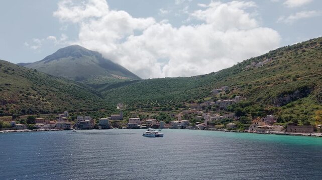 Aerial tracking shot of yachting in the crystalline turquoise waters of Limeni village on Mani Peninsula with mountains in the backdrop under cloudy sky