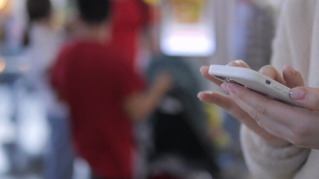 Close-up of hands holding smartphone. Background shows blurred people in public space. Focus on device use, ignoring surroundings. Ideal for scenes of digital communication or waiting