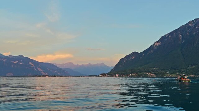 Drone shot of a wakeboard boat cruising across Lake Geneva near Le Bouveret at golden hour, with calm water, alpine mountains, and Swiss lakeside villages.