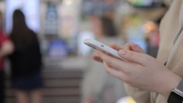 Close-up of woman holding smartphone, fingers scrolling. Blurred background suggests public place like cafe or mall. Hands show focus on digital device. Ideal for scenes of modern communication