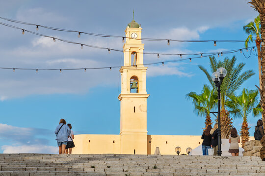 Tourists pause at the top of the wide staircase leading to Kikar Kedumim Square, with sunlight hitting the bell tower of St. Peter's church, in the ancient port of Jaffa, Israel.