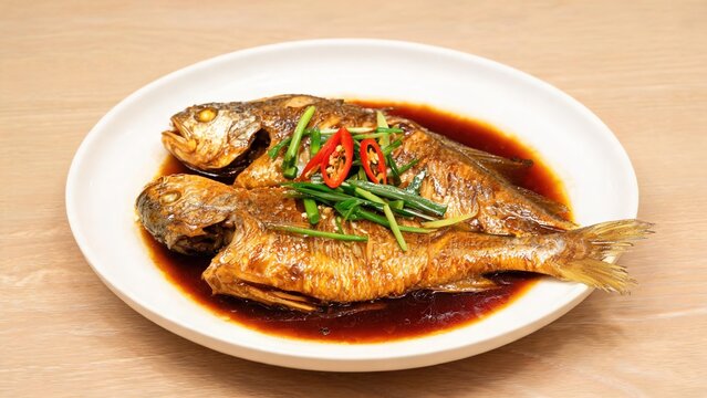 Two Whole Braised Yellow Croakers with Scallions and Red Chili, Classic Homestyle Chinese Braised Seafood Glossy Dark Soy Gravy Served on White Plate Light Wooden Surface Restaurant Food Shot