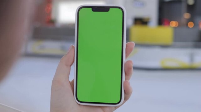 Hand holds phone with green screen. Background shows ice rink with blurred figures. Person focuses on device, intent on digital content. Ideal for apps, ads, or UI mockups