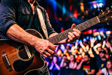 Fototapeta premium Male musician playing acoustic guitar live on stage during concert with cheering crowd in background