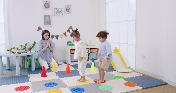 Asian female preschool teacher claps encouragement as preschool girl hops between textured discs, preschool boy steps carefully, indoor daycare cone course, motor skill training, fun exercise