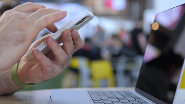Person uses phone while typing on laptop. Hand-focused shot shows multitasking. Background blurred with cafe-like setting. Suitable for tech, work, or communication themes