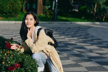 Naklejka premium Woman crouching beside blooming flowers in a park, enjoying a sunny day, casual fashion, backpack ready for a stroll, candid moment of calm and beauty.