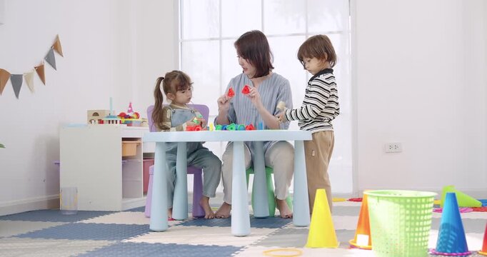 Asian female preschool teacher guiding toddler boy, toddler girl building peg stack toy at low table, bright playroom near window, early childhood education, fine motor skill, color sorting game.
