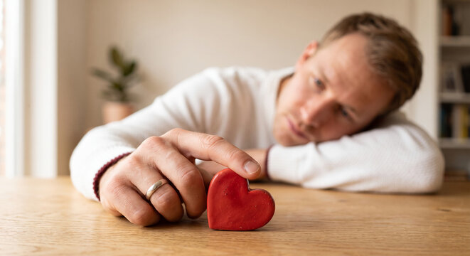 Depressed heartbroken man wearing wedding ring touching a small red heart feeling lonely and sad dealing with divorce grief or relationship problems