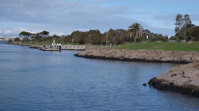 Calm waterfront with rocky seawall, jetty, parkland and people fishing in the distance in Williamstown Melbourne Australia. Scenic coastal reserve. Concept of bayside recreation, coastal living