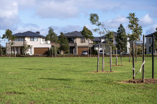 An open public parkland and grass reserve near a row of large, modern double-story family homes in a master-planned residential estate within an outer suburb of Melbourne, Australia. 