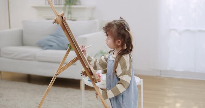 Asian toddler daughter paints on tall easel in living room, steps back, checks artwork, returns brush to palette, continues creative play, calm indoor lifestyle, family bonding weekend home.