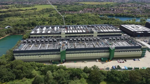 This shows an aerial perspective of a massive data center campus with multiple large buildings. It is situated within a lush green landscape, featuring trees, grass, and nearby water bodies.