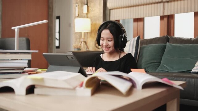 Asian teenage girl greeting in online class, remote learning at home using digital tablet, writing and reading textbooks, self-study for back to school, Homeschooling, university education lifestyle.