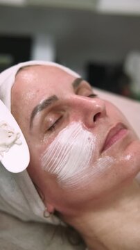 Vertical video. Cream mask. Facial beauty. Esthetician performing professional care applying white product on female skin using brush in salon.