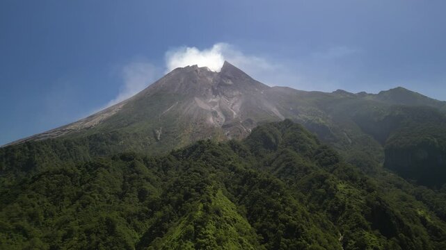 4k aerial drone video footage of Mount Merapi volcano seen from Klangon Hills emitting volcanic smoke from its summit crater, surrounded by forest mountain ridges, clear blue daytime sky.