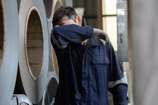 Exhausted male industrial worker wearing blue uniform wiping sweat from face standing near heavy metal coil inside large factory showing extreme tiredness and physical exhaustion from hard manual