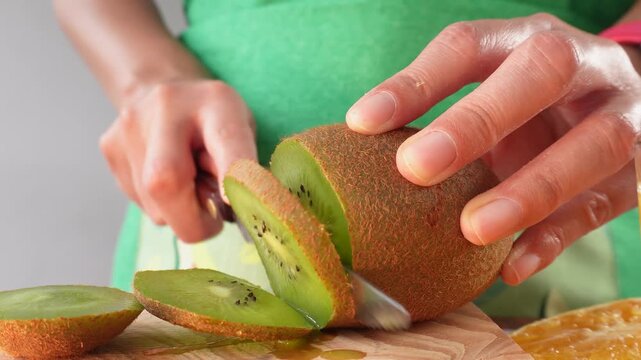 Close-up of hands slicing fresh kiwi fruit on wooden board. Person wears green apron, focused on cutting. Ideal for food preparation