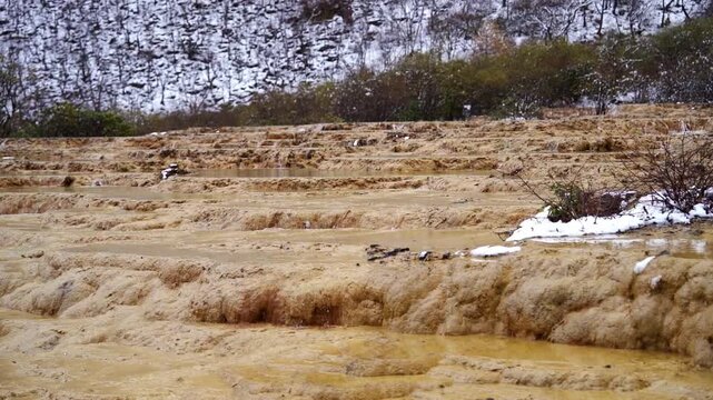 A close-up slow panning shot tracking across the wet, textured ripples of golden limestone travertine terrace steps with patches of fresh white snow.