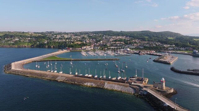 Sailboats line the outer breakwater and marina basins at Howth as a drone flies inward toward the harbour entrance on a clear summer day.