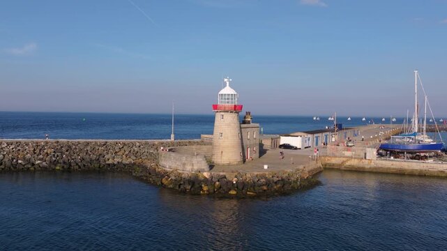 People walk around the stone lighthouse and along the breakwater at Howth Harbour as small sailing boats move across the bay.