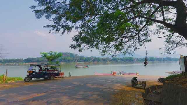 Scenic riverfront with a local tuktuk and scooter under a large tree overlooking the serene town and mountains of Luang Prabang Laos