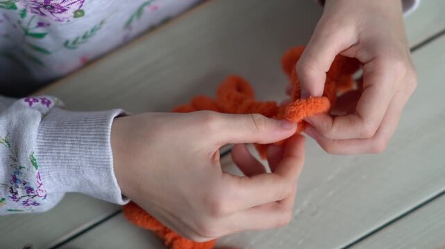Little baby's fingers are knitted using orange plush yarn. Children's hobby. The girl sits holding a woolen thread in her hands.