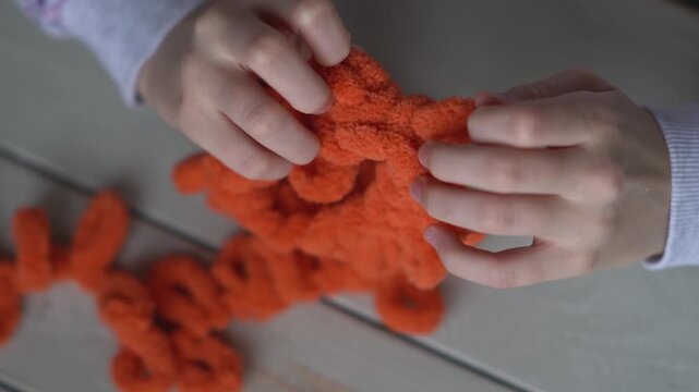 Little baby's fingers are knitted using orange plush yarn.. Children's hobby. The girl sits holding a woolen thread in her hands.