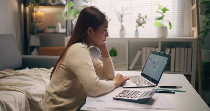 Thoughtful Asian businesswoman with hand on chin looking at laptop screen, analyzing financial chart at home.