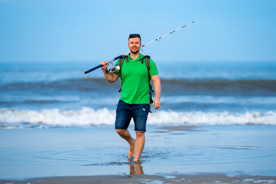 Fishing man enjoys seaside outdoor walk. Fishing rod and basket in hands. Fishing on beach with waves and sand. Fishing lifestyle shows relaxation and hobby.