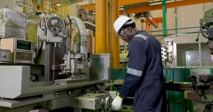 African adult male technician in white hardhat monitors machine table and control area, checking production movement and equipment setting for precision manufacturing in slow motion