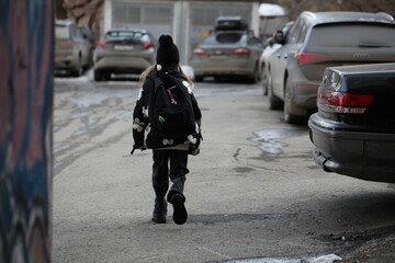 Fototapeta premium Young girl with a school backpack walking away down a city street alley between parked cars, back view.