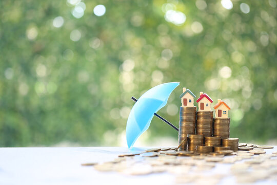 Miniature houses on top of coin stacks protected by a blue umbrella, symbolizing real estate insurance and financial security.