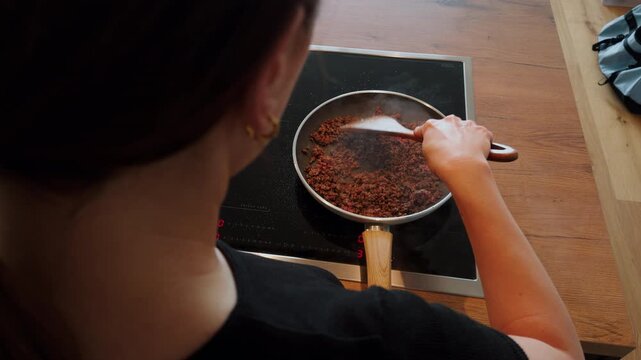 A woman stirs minced beef in a pan on an induction hob while steam rises from the hot mixture. The scene is filmed from above in a modern kitchen.