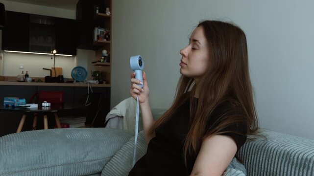 A young woman sits on a sofa at home holding a small handheld fan toward her face. Her eyes are closed as she tries to stay cool in a warm living room.