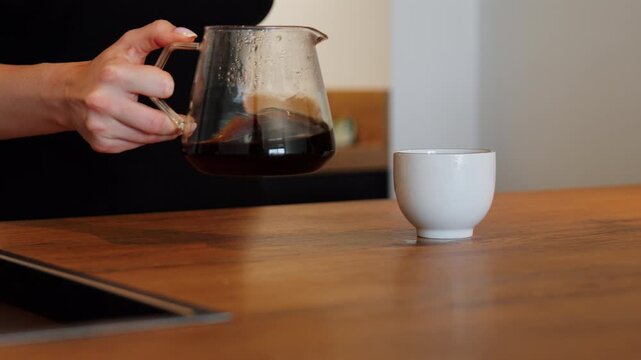 A woman pours freshly brewed filter coffee from a glass server into a white cup on a wooden kitchen counter.