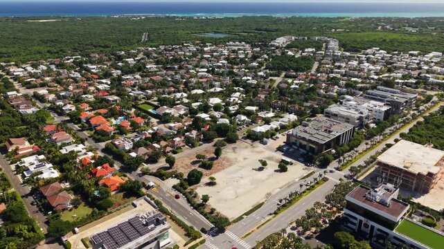 High-angle drone pan over a growing commercial and residential district in Punta Cana Village. Shot shows modern office buildings, ongoing construction, and dense tropical foliage near the Caribbean c