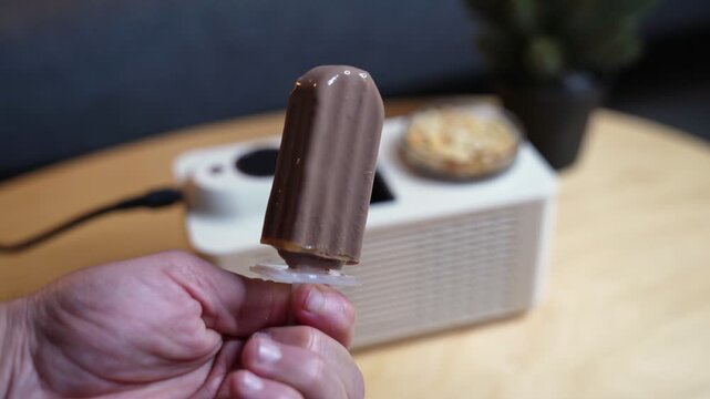 Close-up of a hand holding a freshly made chocolate popsicle. A white mini ice pop maker sits on a wooden table in the blurred background of a brightly lit modern home interior.