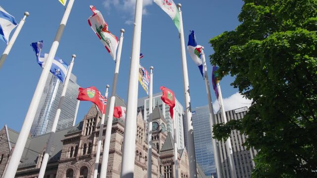 Old Toronto City Hall is gradually obscured as camera moves forward and tilts upward toward international flags waving above the square, Slow Motion