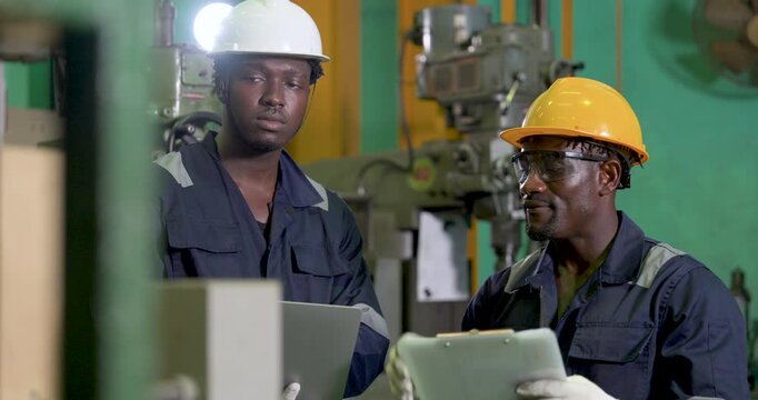 african adult male engineer points across workshop while coworker checks laptop, technical discussion in , safety teamwork, production planning and machine review continue
