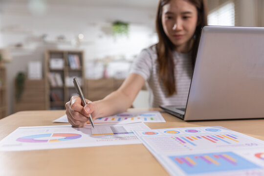 Asian woman working on laptop at desk holding pen over paper chart with attentive expression analyzing business data document in home office environment