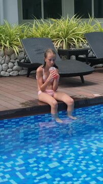 Vertical video. Teenage girl in a pink two-piece swimsuit sitting on the pool deck with her legs dangling in the water, sipping a strawberry iced cocktail beside a resort pool