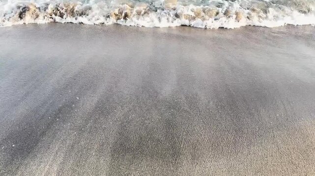 Gentle sea waves roll onto a wide sandy beach, creating soft foam and natural coastal textures in a calm summer seaside scene.