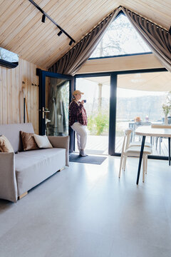 Vertical shot of calm woman drinking coffee inside cozy wooden cabin with lake view