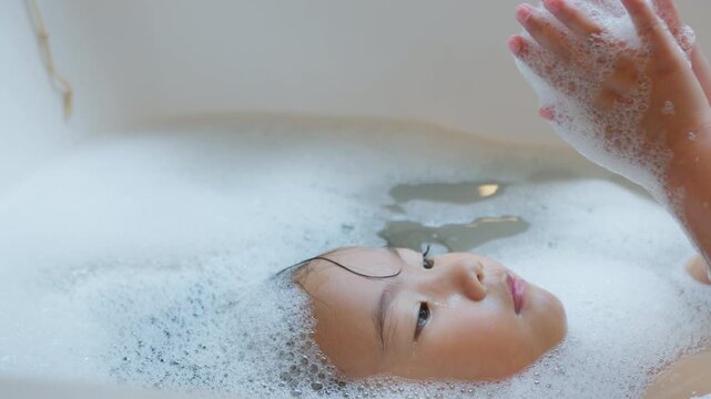A young Asian girl relaxes in a warm bubble bath at home, looking up at her soapy hands while enjoying a moment of sensory play and daily hygiene routine.