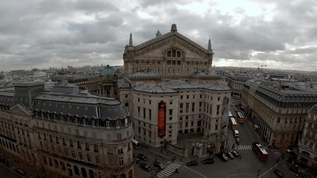 Wide high angle view of the Palais Garnier in Paris, France, showing the historic opera house facade, surrounding streets, traffic, pedestrians and distant Eiffel Tower under a cloudy sky.