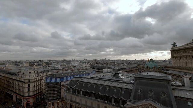 Wide elevated shot overlooking the Palais Garnier opera house and surrounding Haussmannian rooftops in Paris, France, with a moody overcast sky and the city skyline stretching into the distance.
